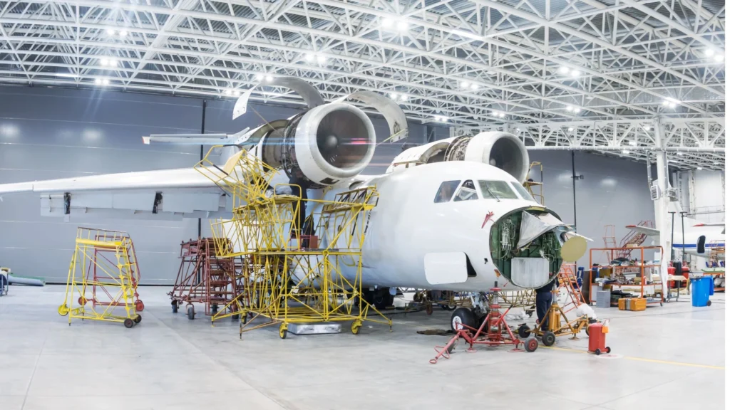 Aircraft undergoing engine and structural maintenance inside an aerospace manufacturing hangar, showing aviation engineering technicians and repair equipment.