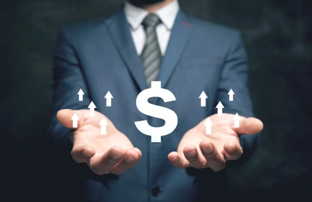 Business professional holding a dollar symbol with upward arrows representing revenue growth and increased profitability.