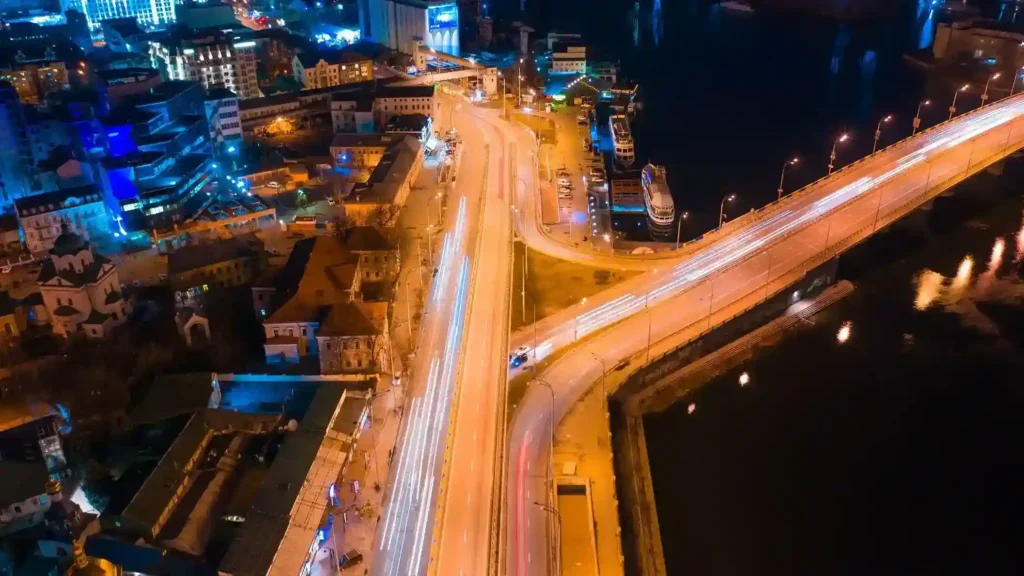 Aerial night view of a modern city bridge with long-exposure traffic light trails reflecting over a dark river, surrounded by illuminated buildings and urban streets.