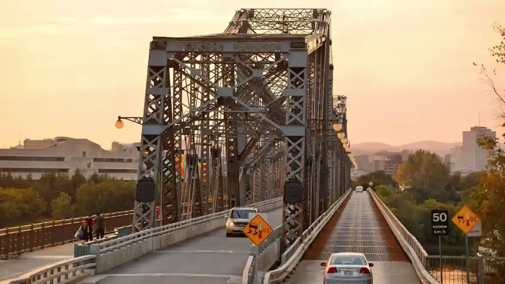 Steel truss bridge during sunset with cars driving across the roadway and city buildings visible in the background.