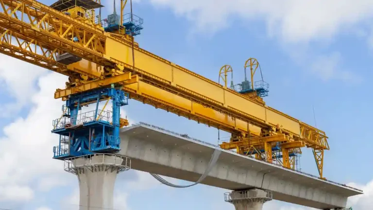 Large yellow industrial gantry crane installing a concrete bridge segment on elevated support pillars under a cloudy sky.
